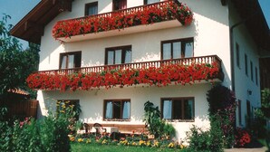 A two-story building with balconies adorned with red flowers, multiple windows, and a garden area with various plants and flowers. - Ferienwohnung "Meine Kleine Auszeit", 50 qm, Dachgeschoss, 1separates Schlafzimmer (Höslwang)