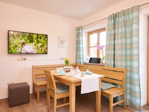 A dining area with a wooden table, four chairs, a bench, a TV mounted on the wall, a window with curtains, and a small plant on the windowsill.