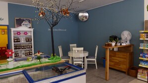 A room with a blue wall, a small round table with chairs, a white shelf with products, a decorative tree, and a fan on a wooden cabinet. - Well-kept apartment in the Eifel. (Lissendorf)