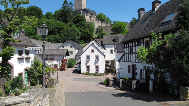 A traditional European village street with stone buildings, a sign reading "Bäckerrei," and a castle in the background. - Well-kept apartment in the Eifel. (Lissendorf)