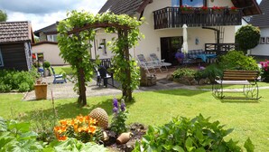 A garden with a pergola, outdoor seating, and a house with a balcony. - Well-kept apartment in the Eifel. (Lissendorf)