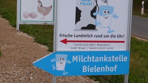 A signboard with text and illustrations, including a chicken and a milk carton character, indicating a local business or service. - Well-kept apartment in the Eifel. (Lissendorf)