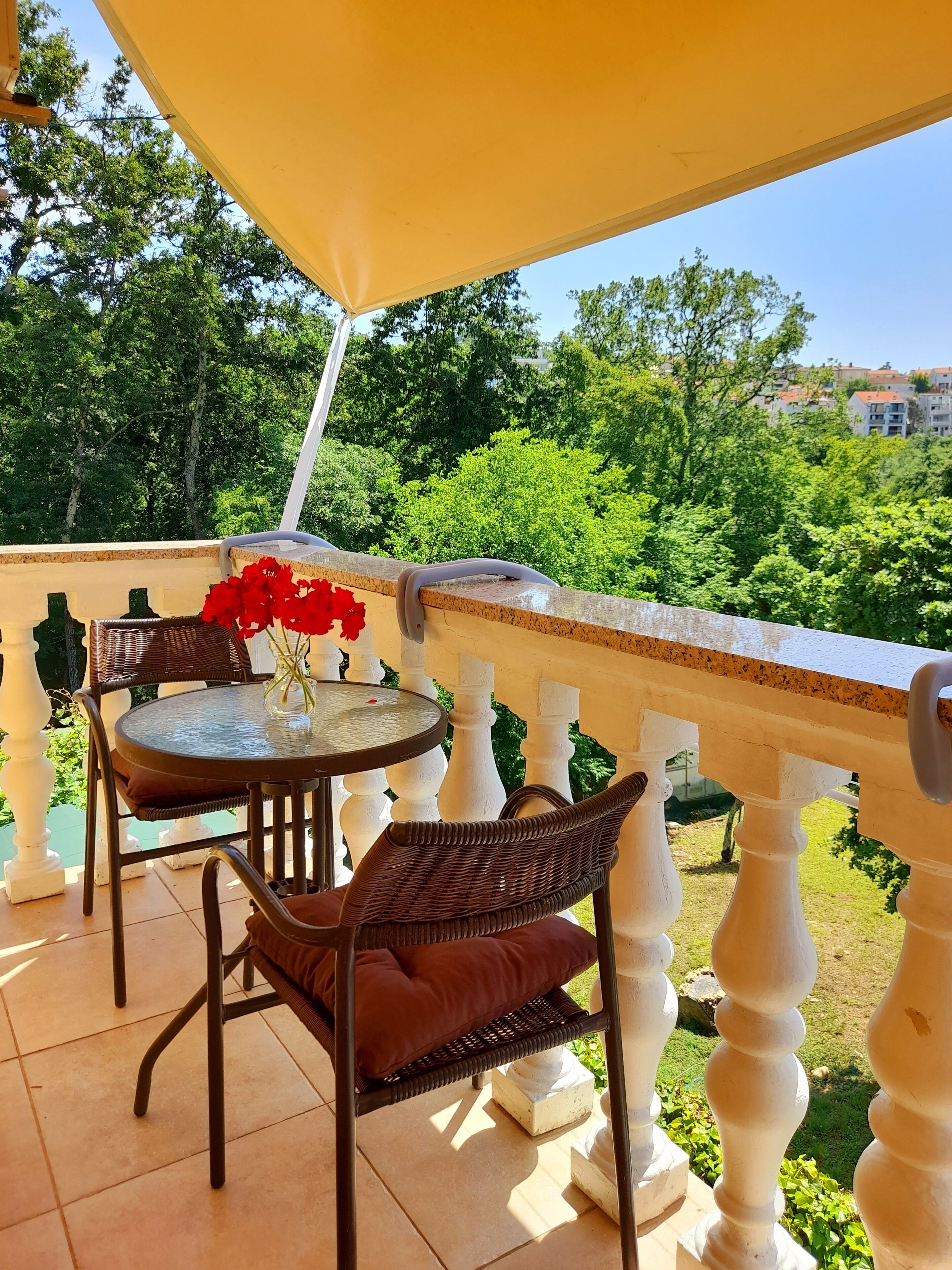 A balcony with a round table and wicker chairs, a yellow umbrella, and a view of greenery and buildings.