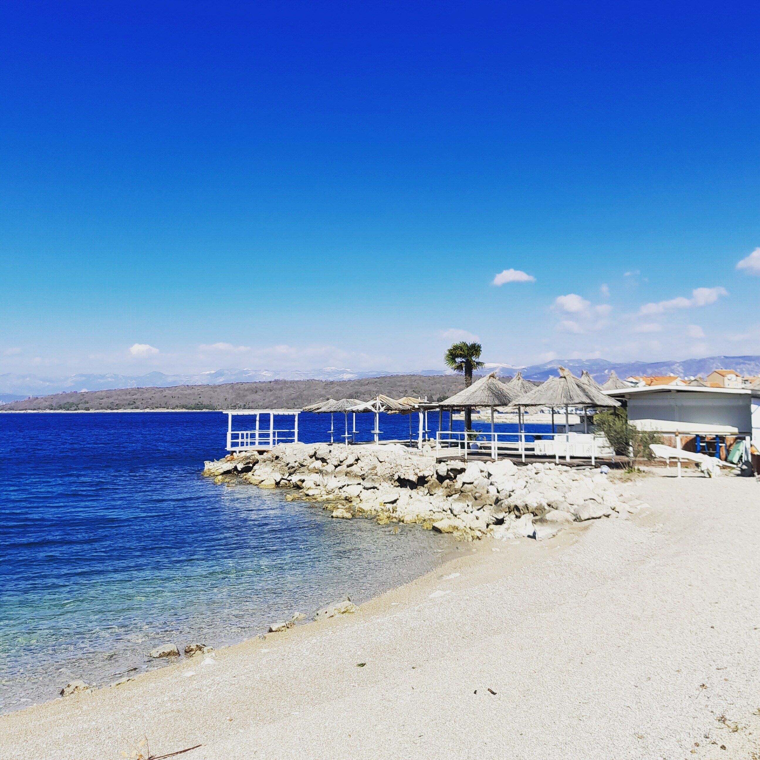 Une plage avec un bâtiment blanc, une construction à toit de chaume et une côte rocheuse.