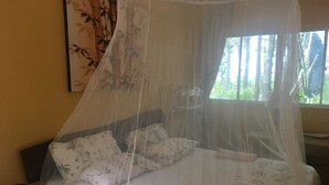 A bed with a mosquito net, a bedside table with a lamp, and a window with a view of greenery. - GreenHouse EcoLodge (Kampung Mantanani)