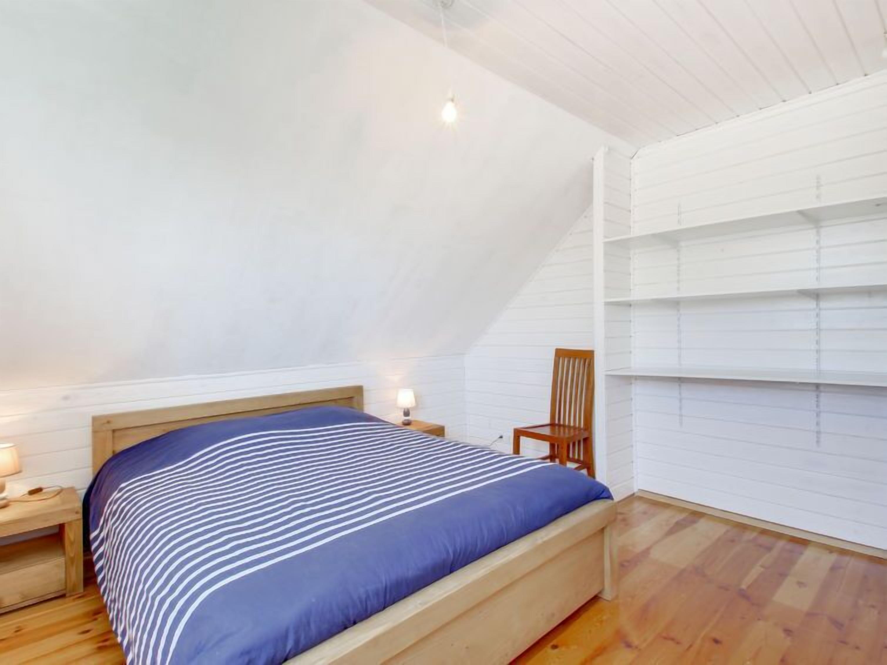 A bedroom with a wooden bed, a nightstand with a lamp, and a chair against a white wall.