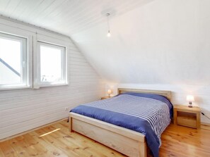 A bedroom with a sloped ceiling, a wooden bed, two bedside tables with lamps, and large windows.