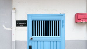 Une porte bleue avec une grille en métal, un paillasson vert à l’entrée et une enseigne sur laquelle on peut lire : « OVER THE SKY ».