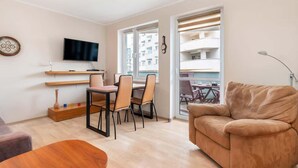 Una sala de estar moderna con un sofá, una mesa de comedor con sillas, un televisor montado en la pared y un balcón con mobiliario exterior.