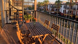 A balcony with a wooden table and chairs, a potted plant, and a view of the street and buildings.