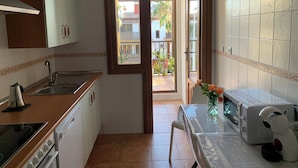 A compact kitchen with a sink, stove, and microwave. A wooden door leads to a balcony with a view of buildings and plants.
