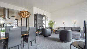 Una sala de estar moderna con comedor, sofás grises, un reposapiés de cuero negro, una lámpara colgante y una cocina al fondo.