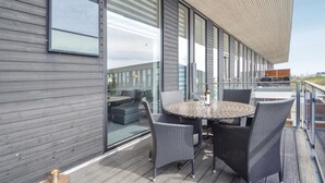 Un área de estar moderna al aire libre con una mesa redonda y sillas de mimbre, ventanas amplias y una terraza de madera.