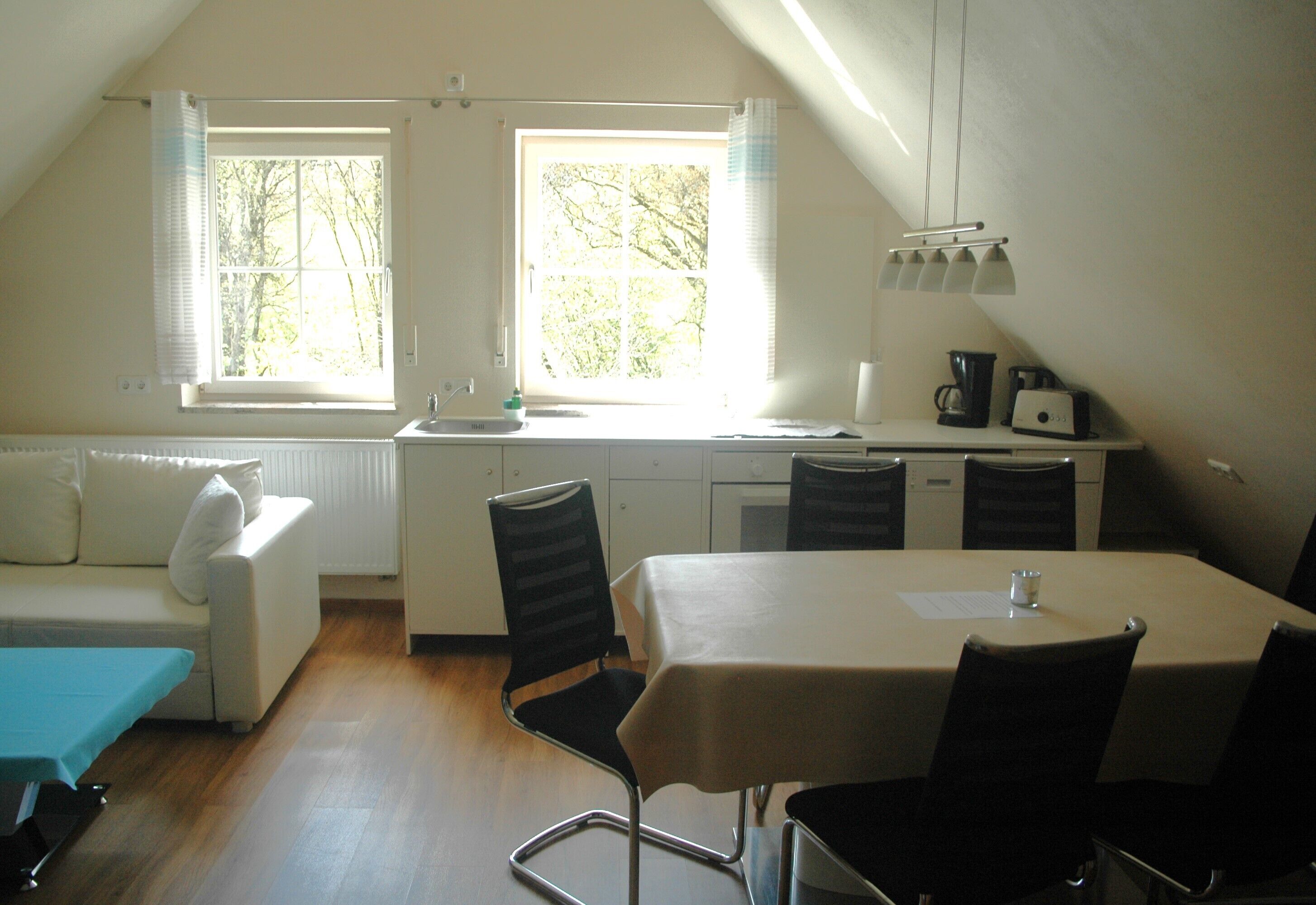 A small, bright room with a dining table, chairs, a sofa, and a window with curtains.