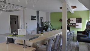 A modern dining area with a long table, chairs, and a kitchen in the background. - Vacation home/event location, 285 m² living area, 300 m² secluded outdoor area  (Rieneck)