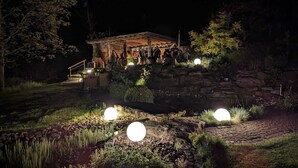 A rustic outdoor pavilion with stone walls and a wooden roof, surrounded by a landscaped garden with rocks and plants, and illuminated by spherical lights. - Vacation home/event location, 285 m² living area, 300 m² secluded outdoor area  (Rieneck)