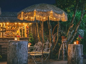 An outdoor bar area with a thatched umbrella, wooden furniture, and a bar stocked with bottles.