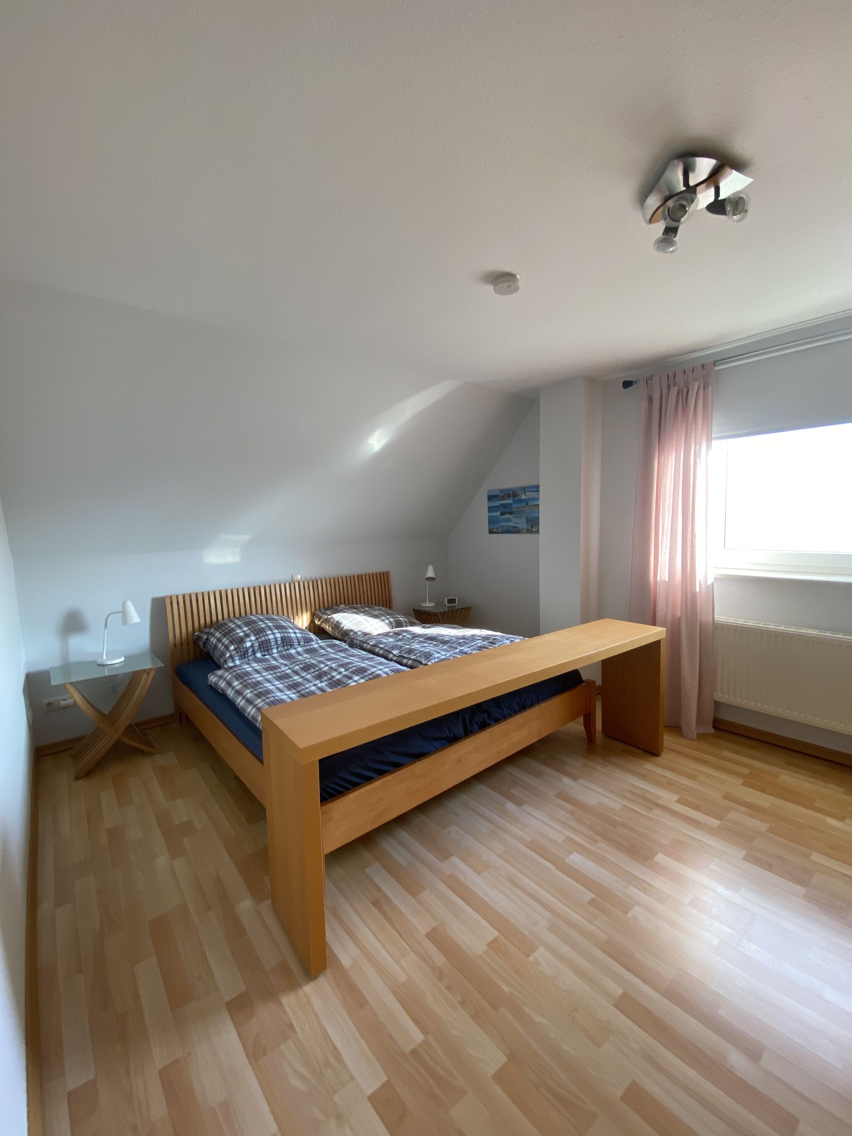 A bedroom with a sloped ceiling, a wooden bed frame, a desk with a lamp, and a window with curtains.