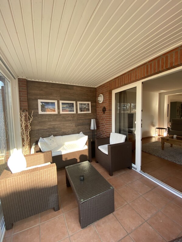 A sunlit patio with wicker furniture, a glass door leading to an indoor area, and a brick wall with framed pictures.