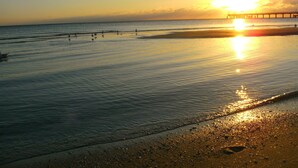 Eine Strandansicht mit einem Steg, der sich in ruhiges Wasser erstreckt, in dem der Sonnenuntergang widergespiegelt wird.