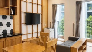 A modern living room with a wooden dining table, chairs, a sofa, a TV mounted on a wooden panel, and a balcony with a view. - Minh Hưng Apartment (Da Nang)