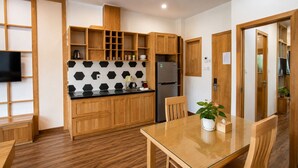 A compact kitchen with wooden cabinets, a black and white tiled backsplash, a stainless steel refrigerator, and a dining table with chairs. - Minh Hưng Apartment (Da Nang)