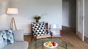 A living room with a sofa, a chair with a geometric pillow, a glass coffee table with fruit, a lamp, and a bookshelf. - Modern flat facing south with view over mountains and lake (Murnau)