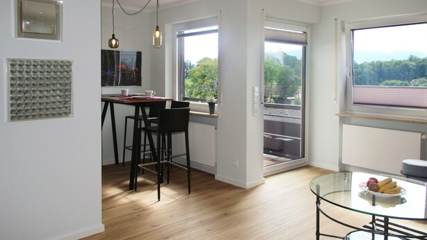 A modern kitchen with a dining area, large windows with a view, and a glass table with fruit. - Modern flat facing south with view over mountains and lake (Murnau)