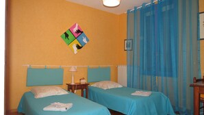 Ein Zimmer mit zwei Betten, einem Nachttisch mit Lampe, einem kleinen Tisch und einem farbenfrohen Kunstwerk an der Wand.