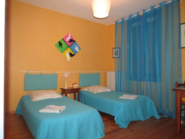 Una habitación con dos camas, una mesita de noche con lámpara, una pequeña mesa y una obra de arte colorida en la pared.