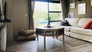 A living room with a white sofa, a wooden coffee table, a potted plant, and a large window with curtains.