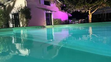 Une piscine à l’eau claire, une maison dont l’extérieur est illuminé de lumière violette, et un arbre en arrière-plan.