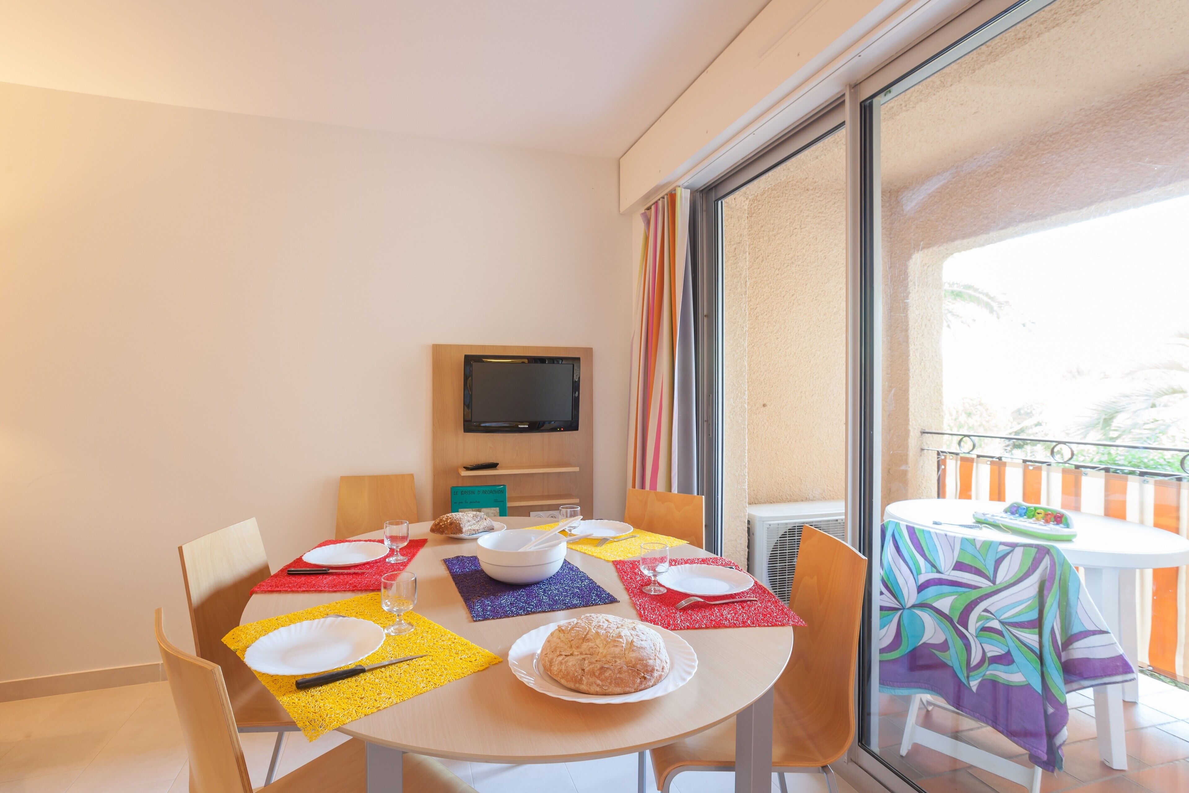 A dining area with a table set for four, a television above the table, and a balcony with a view.