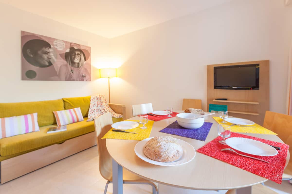 Un coin repas avec une table dressée pour deux personnes, un canapé, une télévision et une lampe.
