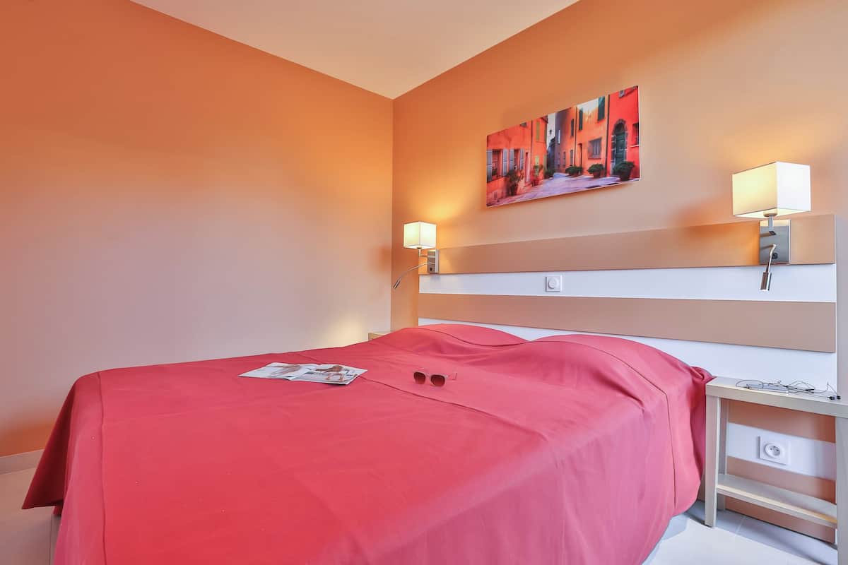 Une chambre d’hôtel avec un lit recouvert d’une couverture rouge, une table de chevet avec une lampe et un tableau accroché au mur.