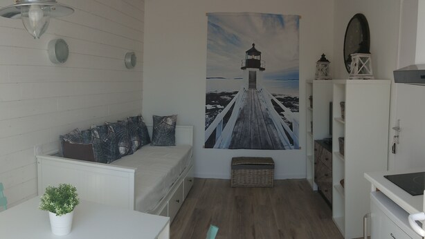 A room with a white bed, a white table with a potted plant, a wooden floor, a painting of a lighthouse, and a white cabinet. - Our islands : Ile du Pilier - 200m Beach (Saint-Jean-de-Monts)