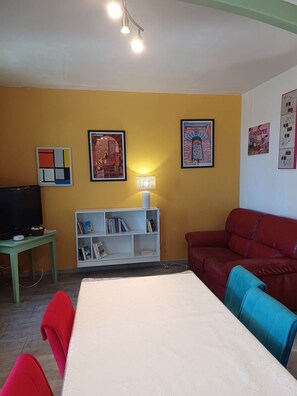 Un salon avec un mur jaune, un canapé rouge, une chaise bleue, une table blanche et un téléviseur posé sur un meuble vert.
