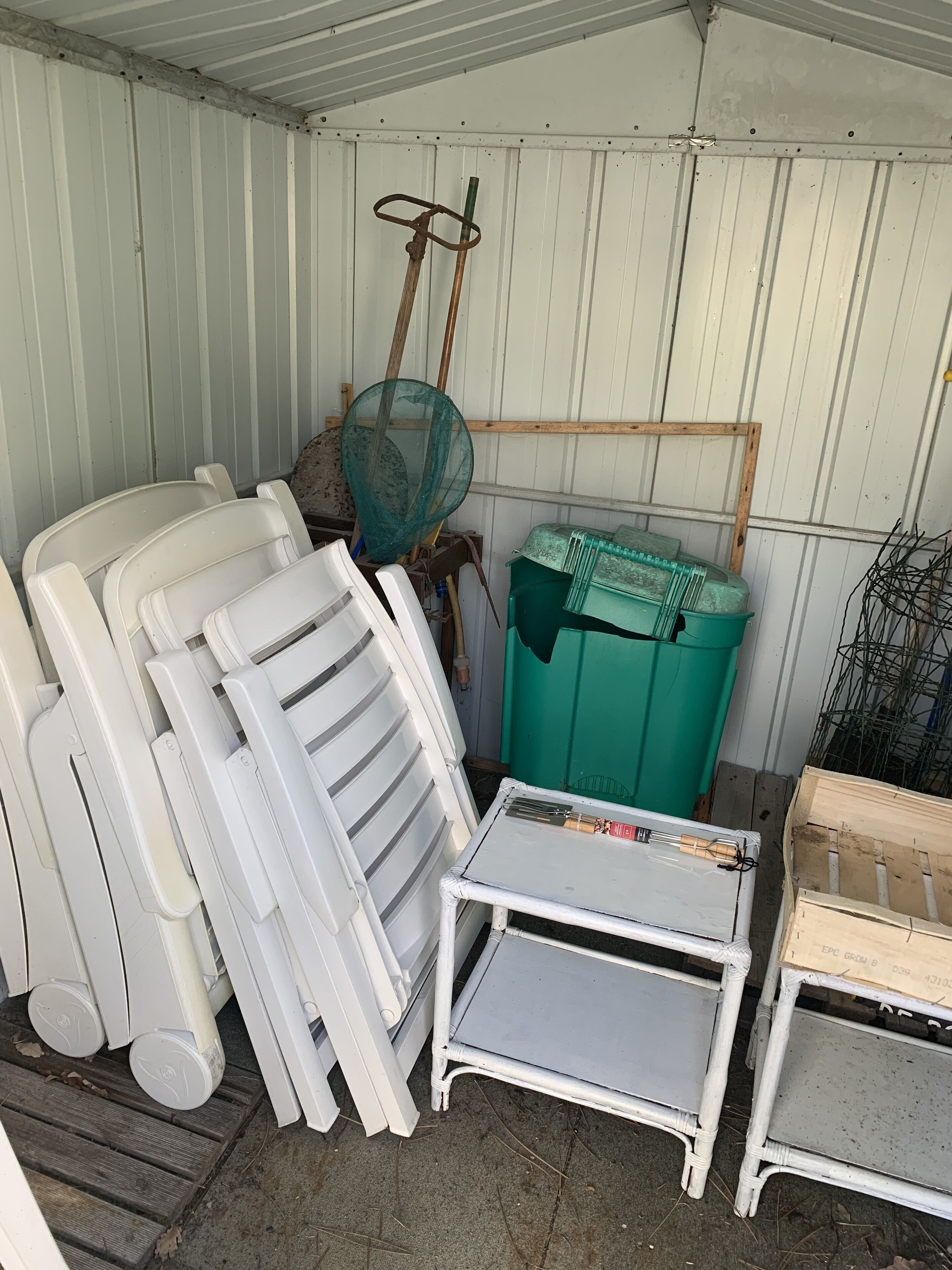 A storage area with white plastic chairs, a metal table, and a green trash bin.