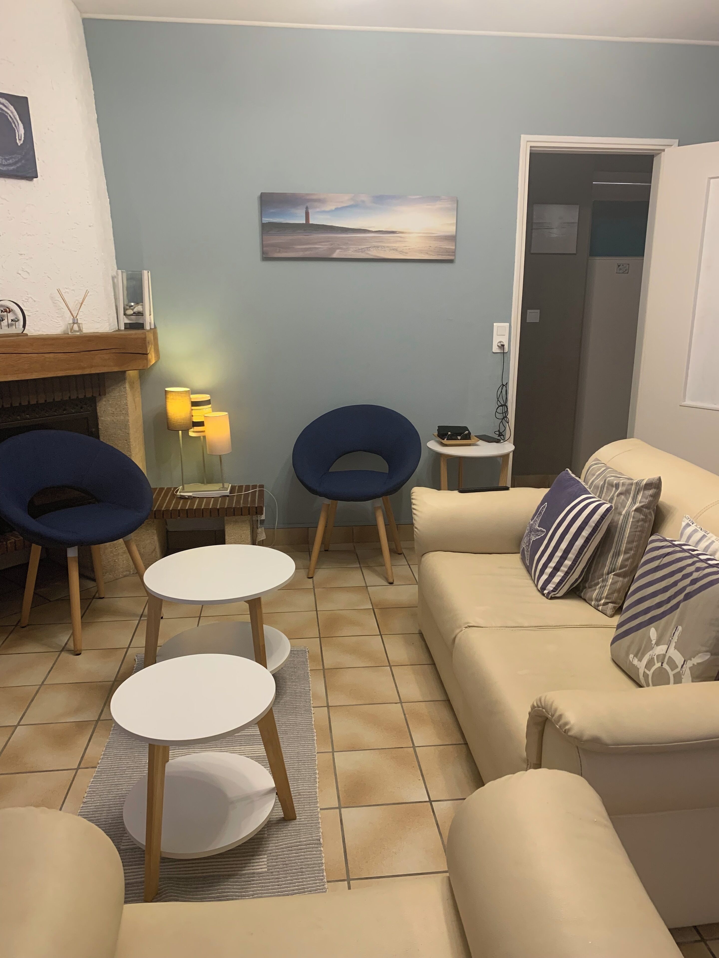 Un salon avec un canapé beige, deux fauteuils bleus, une petite table blanche et une cheminée.