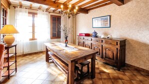 Une table à manger en bois, surmontée d’un lustre, un buffet avec des tiroirs et une fenêtre avec des rideaux.