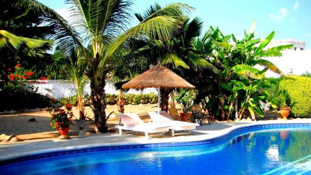 A swimming pool with lounge chairs and a thatched umbrella, surrounded by palm trees and tropical plants. - Villa pirogue: The huts of Saly (M bour)