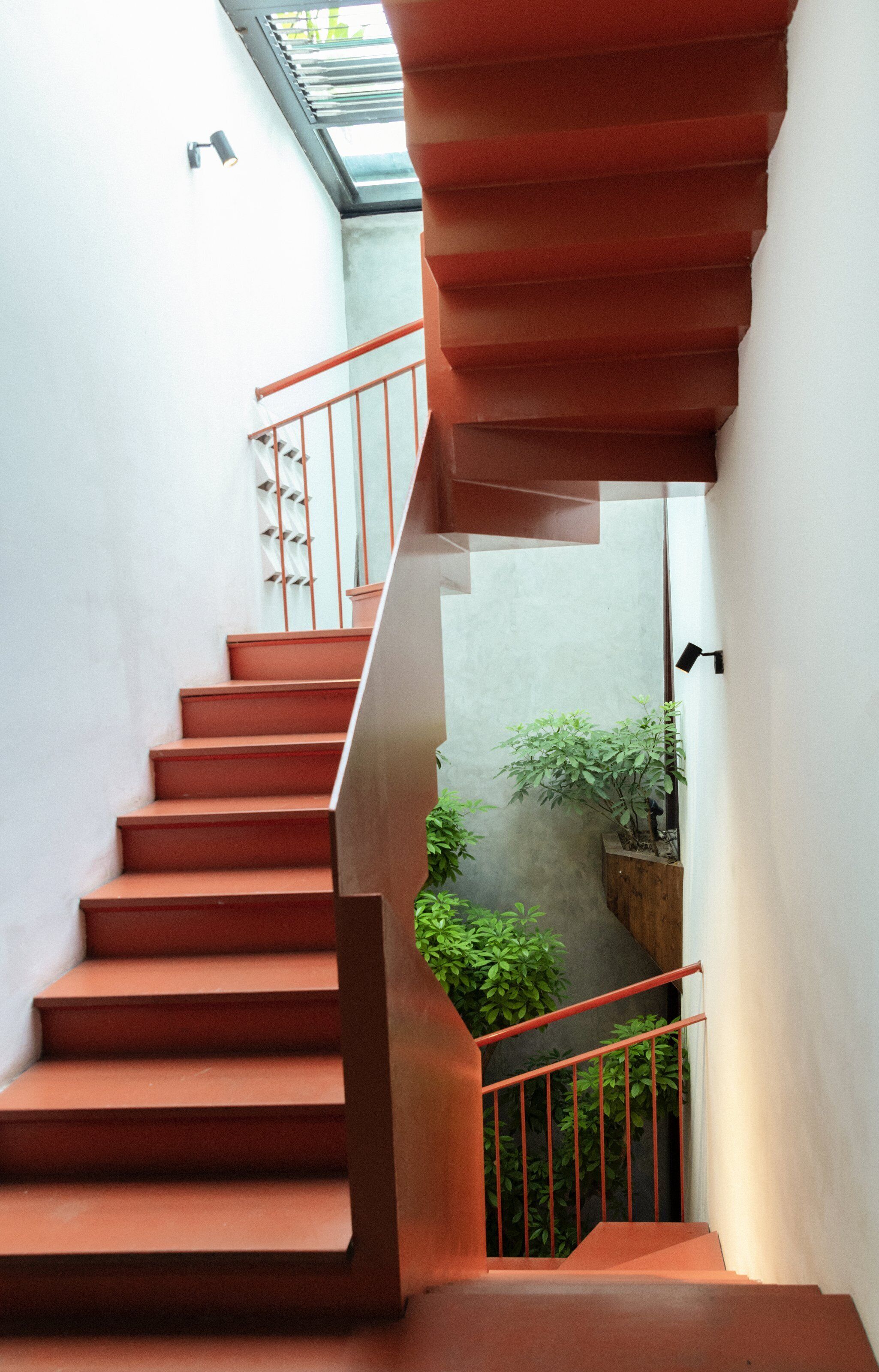 A spiral staircase with red steps and metal railings, leading to a landing with a view of greenery.