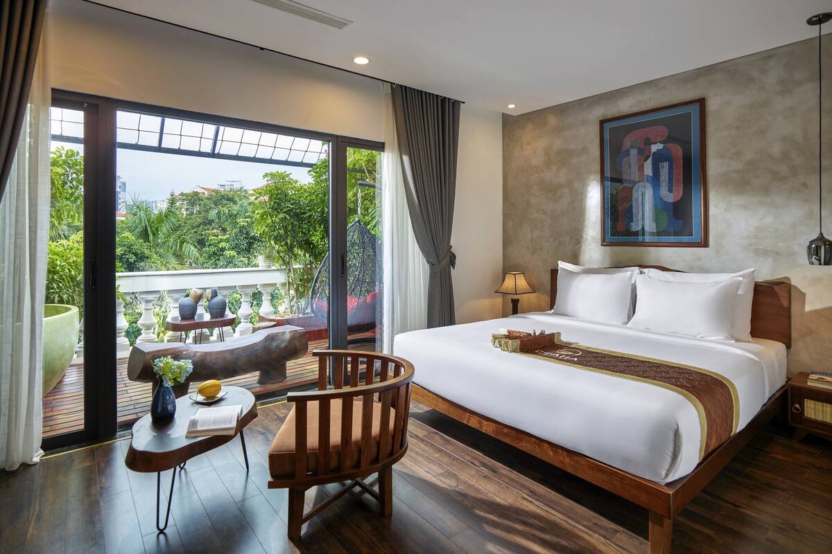 A hotel room with a large bed, a wooden chair, a small table with a vase and book, and a view of greenery through large windows.