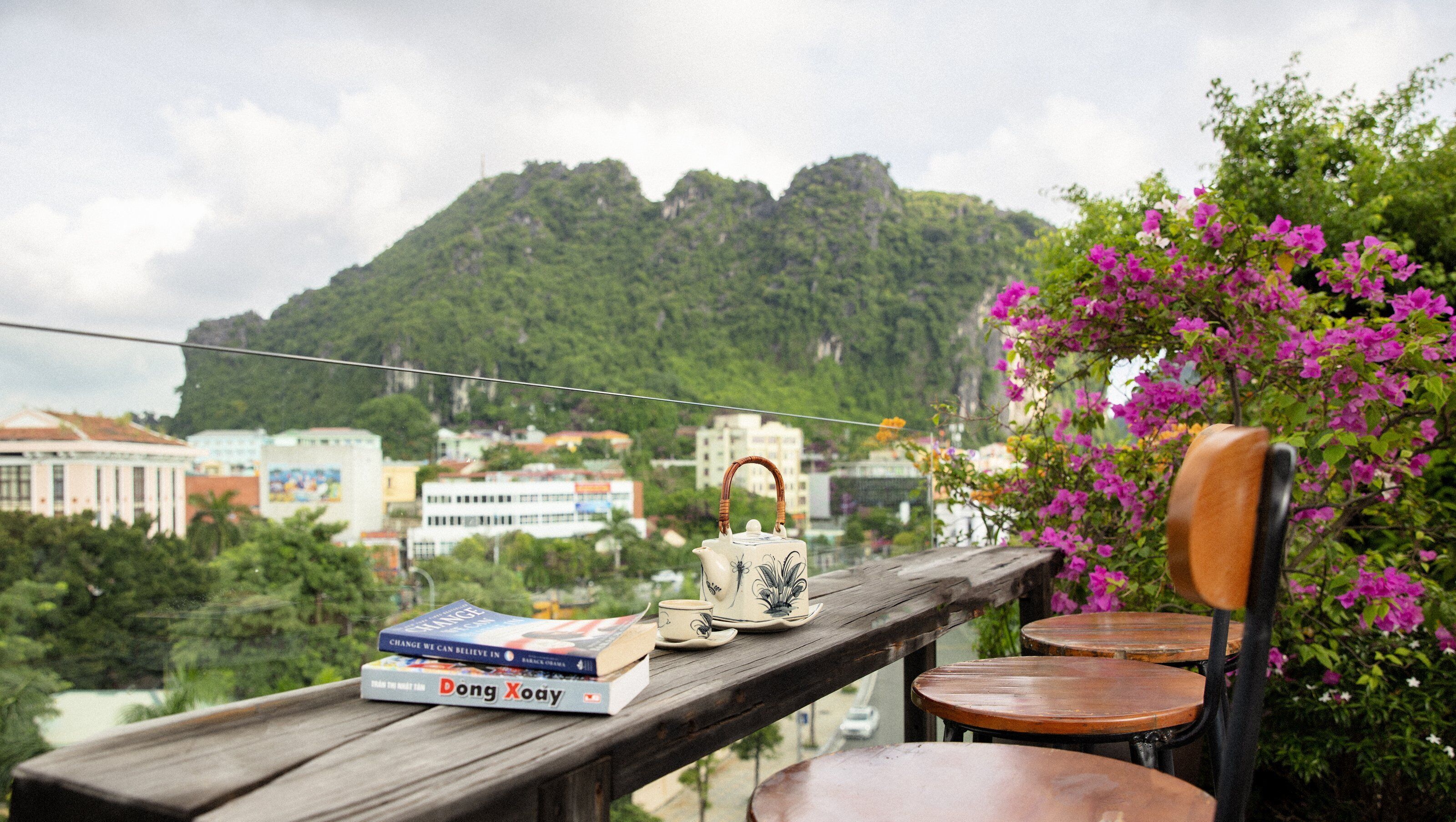 A wooden table with a teapot and cups, a chair, and books on a balcony overlooking a city with mountains in the background.