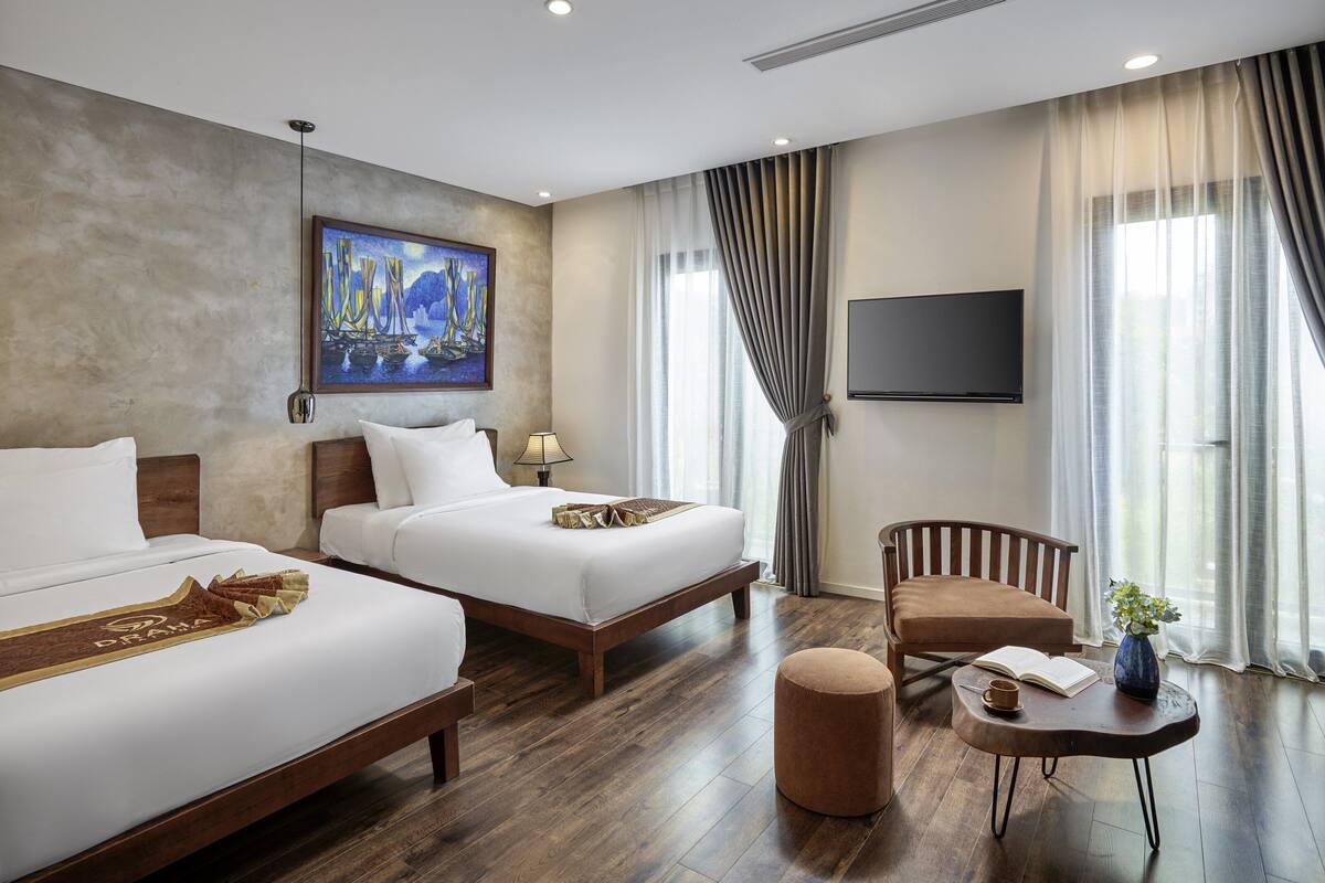 A hotel room with two beds, a TV, a painting, a chair, and a table with a book and a vase.