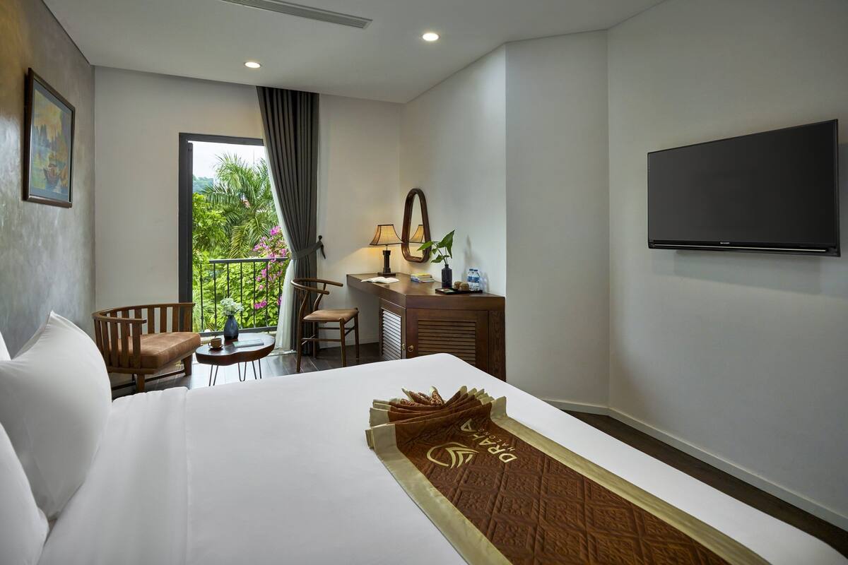 A hotel room with a bed, a desk with a lamp, a TV, and a view of greenery through the window.