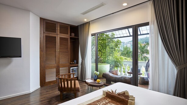 Une chambre d’hôtel comprenant un lit, une télévision, un coin salon avec une table et des chaises, et un balcon agrémenté de plantes.