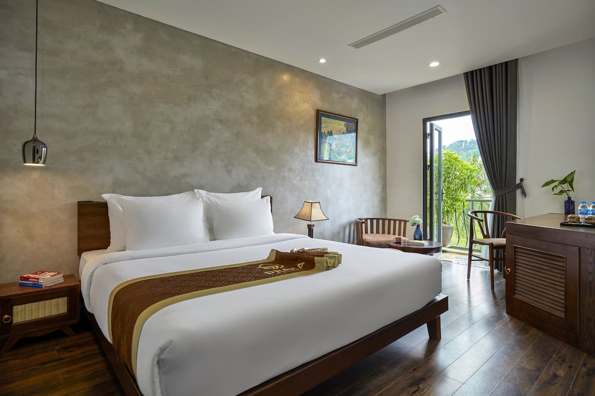 A hotel room with a large bed, a nightstand, a lamp, a chair, and a view of greenery through a window.