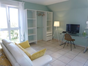 A room with a white sofa, a glass desk with a chair, a TV, and a wardrobe with hanging clothes.