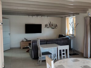Una sala de estar moderna con un sofá seccional gris, una mesa de comedor blanca con sillas, un televisor de pantalla plana montado en la pared y una lámpara de pie.